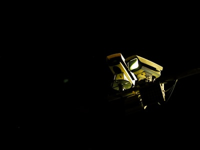 A pair of surveillance cameras is mounted on a pole, illuminated against a dark background. The cameras have rectangular shapes with distinct lenses and are positioned to cover different angles.