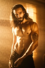 A muscular male figure stands shirtless, illuminated by warm, golden light filtering through window blinds. He has long hair and a beard, showcasing a tattoo on his chest and another tattoo on his arm. The man holds a knife in his hand, positioned near the waistband of his pants.