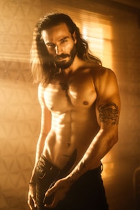 A muscular male figure stands shirtless, illuminated by warm, golden light filtering through window blinds. He has long hair and a beard, showcasing a tattoo on his chest and another tattoo on his arm. The man holds a knife in his hand, positioned near the waistband of his pants.