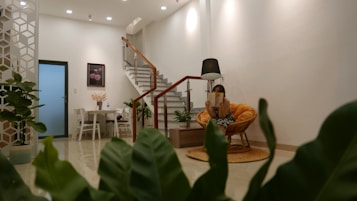 A modern, cozy living space with an open layout decorated with potted plants. A small round dining table with chairs is located near a white door, and a staircase with a wooden handrail leads upward. A woman sits in an orange cushioned chair, reading under a black floor lamp. The room is well-lit with ceiling lights, and a framed picture with flowers hangs on the wall.