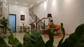 A modern, cozy living space with an open layout decorated with potted plants. A small round dining table with chairs is located near a white door, and a staircase with a wooden handrail leads upward. A woman sits in an orange cushioned chair, reading under a black floor lamp. The room is well-lit with ceiling lights, and a framed picture with flowers hangs on the wall.
