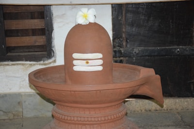 A traditional earthen lingam with white horizontal stripes sits in a decorative basin. A white flower is placed on top, and it's set within an indoor setting featuring rustic walls and wooden accents.