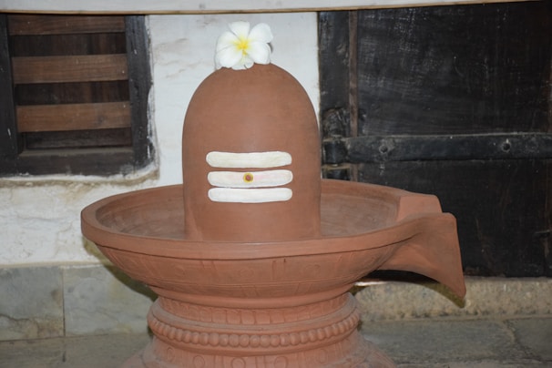 A traditional earthen lingam with white horizontal stripes sits in a decorative basin. A white flower is placed on top, and it's set within an indoor setting featuring rustic walls and wooden accents.