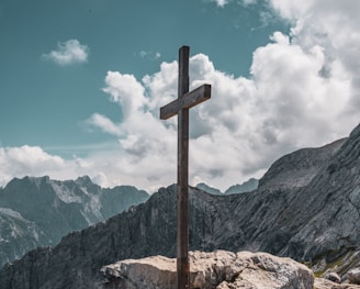 brown wooden cross on brown rock near green mountains under blue sky and white clouds during