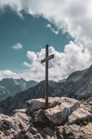 brown wooden cross on brown rock near green mountains under blue sky and white clouds during
