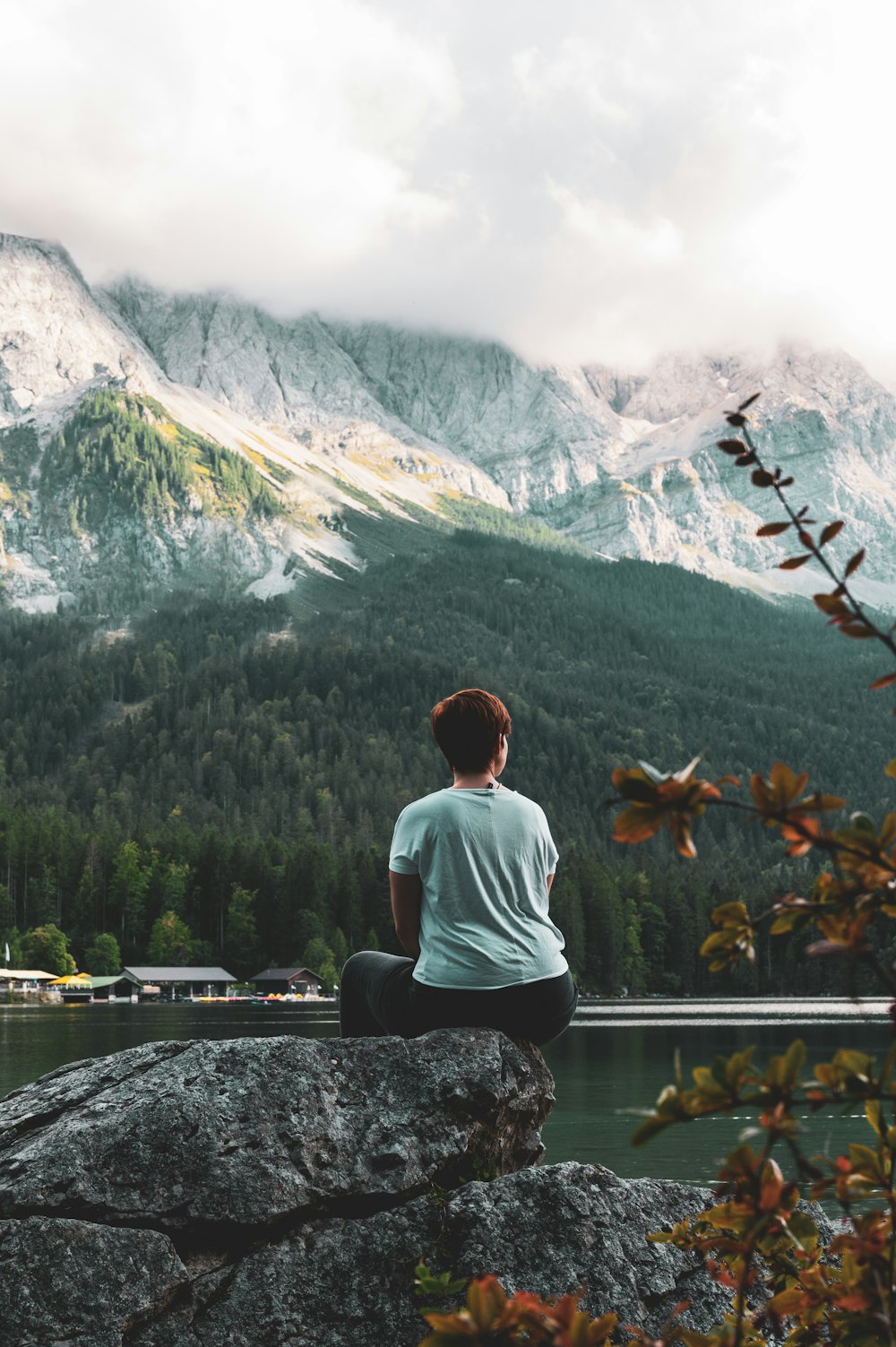 man in white shirt sitting on rock near lake and mountains during daytime