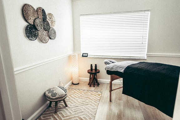 A cozy room with a massage table covered by a dark blanket, a stool with a patterned cushion, a woven wall art decoration, a small table with two bottles, and a tall lamp beside the table. Natural light filters through the closed blinds on the window, and the room has a calm, spa-like ambiance.