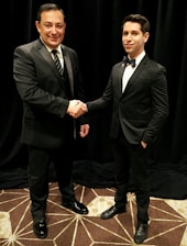 Two men dressed in formal black suits are shaking hands in an indoor setting with a dark curtain backdrop. The man on the left is holding a piece of paper, and both have a professional demeanor.