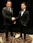 Two men dressed in formal black suits are shaking hands in an indoor setting with a dark curtain backdrop. The man on the left is holding a piece of paper, and both have a professional demeanor.