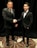 Two men dressed in formal black suits are shaking hands in an indoor setting with a dark curtain backdrop. The man on the left is holding a piece of paper, and both have a professional demeanor.