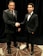 Two men dressed in formal black suits are shaking hands in an indoor setting with a dark curtain backdrop. The man on the left is holding a piece of paper, and both have a professional demeanor.