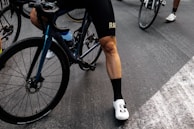 A cyclist wearing black cycling shorts and white cycling shoes is standing beside a blue and black bicycle. The focus is on the cyclist's legs and the bike's rear wheel. The scene takes place on a paved road with several other cyclists visible in the background.