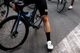 A cyclist wearing black cycling shorts and white cycling shoes is standing beside a blue and black bicycle. The focus is on the cyclist's legs and the bike's rear wheel. The scene takes place on a paved road with several other cyclists visible in the background.