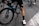 A cyclist wearing black cycling shorts and white cycling shoes is standing beside a blue and black bicycle. The focus is on the cyclist's legs and the bike's rear wheel. The scene takes place on a paved road with several other cyclists visible in the background.