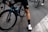 A cyclist wearing black cycling shorts and white cycling shoes is standing beside a blue and black bicycle. The focus is on the cyclist's legs and the bike's rear wheel. The scene takes place on a paved road with several other cyclists visible in the background.
