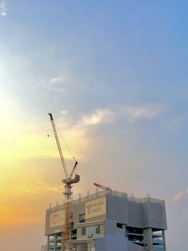 A construction site with a tall crane towering over a partially built structure against a backdrop of a vibrant sky transitioning from warm yellow to a soft blue. The building displays some branding, and the crane is prominently positioned on the left side.