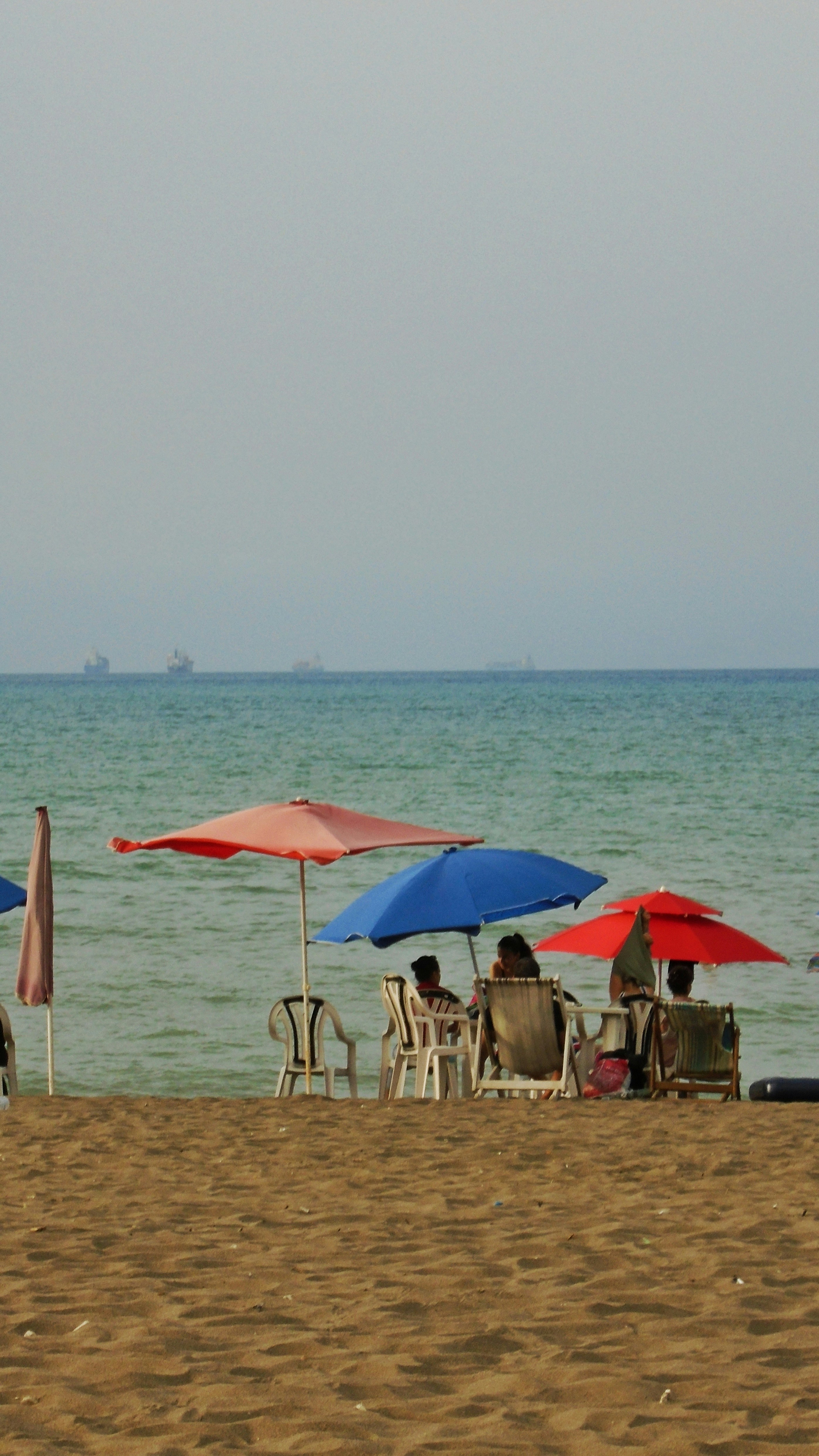 Beach photograph showing colorful umbrellas and lounge chairs on the sand with a calm sea and distant ships on the horizon.