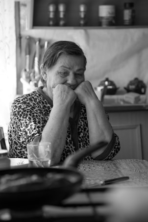 An elderly woman with a thoughtful expression sits at a table in a domestic kitchen setting. She rests her chin on her hands, wearing a patterned garment. Various kitchen items, including a glass, a frying pan, and utensils, can be seen on the table.