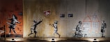 The image features a series of graffiti art on a wall. On the left, a girl is hula hooping. In the center, two children are playing, reaching towards a floating sign that reads 'No Ball Games'. On the right, a child is drawing a house while another child holds wooden planks, with flowers drawn below them.