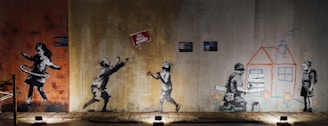 The image features a series of graffiti art on a wall. On the left, a girl is hula hooping. In the center, two children are playing, reaching towards a floating sign that reads 'No Ball Games'. On the right, a child is drawing a house while another child holds wooden planks, with flowers drawn below them.