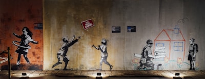 The image features a series of graffiti art on a wall. On the left, a girl is hula hooping. In the center, two children are playing, reaching towards a floating sign that reads 'No Ball Games'. On the right, a child is drawing a house while another child holds wooden planks, with flowers drawn below them.