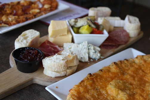 A wooden board is artfully arranged with a selection of cheeses including chunks of cheddar and blue cheese, slices of cured meats, various types of bread, and a small dish of jam. A bowl containing pickled vegetables is also visible. Nearby, there is a plate with a dish that appears to be a type of cheese-filled pastry or pizza.