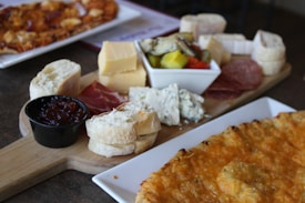A wooden board is artfully arranged with a selection of cheeses including chunks of cheddar and blue cheese, slices of cured meats, various types of bread, and a small dish of jam. A bowl containing pickled vegetables is also visible. Nearby, there is a plate with a dish that appears to be a type of cheese-filled pastry or pizza.