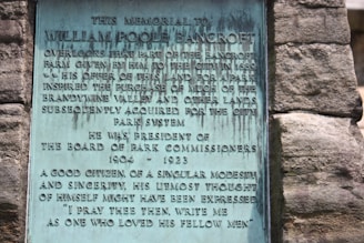 A plaque mounted on a stone wall honors William Poole Bancroft, highlighting his contributions to parkland donations and his role as President of the Board of Park Commissioners from 1904 to 1923. The text describes his character as modest and sincere.
