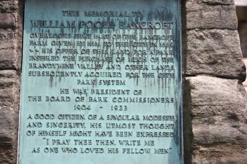A plaque mounted on a stone wall honors William Poole Bancroft, highlighting his contributions to parkland donations and his role as President of the Board of Park Commissioners from 1904 to 1923. The text describes his character as modest and sincere.