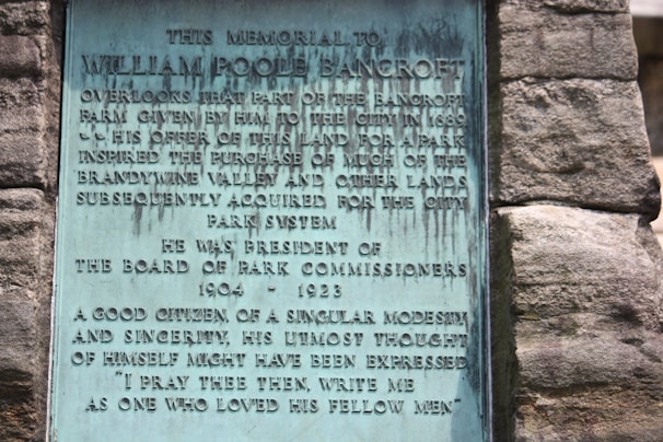 A plaque mounted on a stone wall honors William Poole Bancroft, highlighting his contributions to parkland donations and his role as President of the Board of Park Commissioners from 1904 to 1923. The text describes his character as modest and sincere.