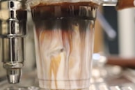 A close-up view of a glass filled with a swirling mixture of dark coffee and white milk. The colors blend beautifully, creating a marbled effect. The liquid is being poured from a metallic espresso machine component, visible to the side. The background is blurred, placing emphasis on the glass and the enticing mixture within.