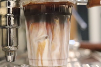 A close-up view of a glass filled with a swirling mixture of dark coffee and white milk. The colors blend beautifully, creating a marbled effect. The liquid is being poured from a metallic espresso machine component, visible to the side. The background is blurred, placing emphasis on the glass and the enticing mixture within.