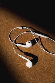 White earphones with a wire rest on a brown textured surface, partially illuminated by light from the left side, creating strong shadows.
