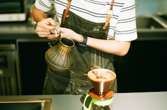 woman pouring brown liquid on clear glass mug