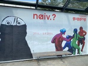 A large, outdoor advertisement features artwork for an exhibition titled 'naiv.?' at museum gugging. On the left, there's a monochrome, sketch-like figure with wide eyes and a partially obscured face. On the right, vivid, colorful human figures are interacting in a dynamic, almost playful manner. Text details the dates and type of the exhibition, described as 'naive kunst aus der sammlung infeld', running from January 27 to September 5, 2021. A simple wooden bench is situated in front of the advertisement.
