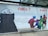 A large, outdoor advertisement features artwork for an exhibition titled 'naiv.?' at museum gugging. On the left, there's a monochrome, sketch-like figure with wide eyes and a partially obscured face. On the right, vivid, colorful human figures are interacting in a dynamic, almost playful manner. Text details the dates and type of the exhibition, described as 'naive kunst aus der sammlung infeld', running from January 27 to September 5, 2021. A simple wooden bench is situated in front of the advertisement.