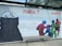 A large, outdoor advertisement features artwork for an exhibition titled 'naiv.?' at museum gugging. On the left, there's a monochrome, sketch-like figure with wide eyes and a partially obscured face. On the right, vivid, colorful human figures are interacting in a dynamic, almost playful manner. Text details the dates and type of the exhibition, described as 'naive kunst aus der sammlung infeld', running from January 27 to September 5, 2021. A simple wooden bench is situated in front of the advertisement.