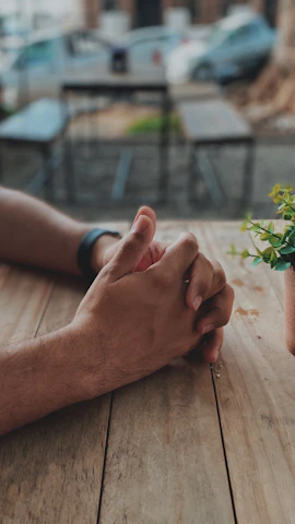 Close-up of hands resting gently on a wooden table with a small potted plant nearby, symbolizing grounding.