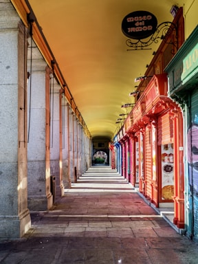 A narrow, covered walkway with colorful shopfronts on one side and stone columns on the other, casting long shadows across the stone pavement. The ceiling is painted a warm yellow, adding to the vibrant atmosphere. Signage in Spanish, including 'Museo del Jamon,' is visible, suggesting a culinary theme.