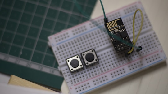 A breadboard setup featuring an electronic circuit. Two tactile push buttons and an ESP8266 module are connected with several jumper wires. The breadboard rests on a surface with a green cutting mat and a notepad visible in the background.