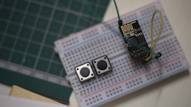 A breadboard setup featuring an electronic circuit. Two tactile push buttons and an ESP8266 module are connected with several jumper wires. The breadboard rests on a surface with a green cutting mat and a notepad visible in the background.