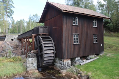 A rustic wooden watermill with a large vertical wheel, located by a small stream. The structure is surrounded by lush green vegetation and trees, creating a serene natural setting. The building has a classic, traditional design with small paned windows and a tiled roof.