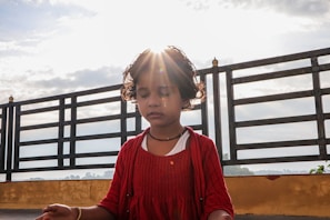 A young child with closed eyes, dressed in a red outfit, sits outdoors with a bright light shining from behind, creating a halo effect. The child appears to be meditating on a balcony or terrace with a metal railing and cloudy sky in the background.