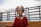 A young child with closed eyes, dressed in a red outfit, sits outdoors with a bright light shining from behind, creating a halo effect. The child appears to be meditating on a balcony or terrace with a metal railing and cloudy sky in the background.