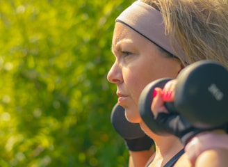A woman with a headband and blonde hair holds a pair of dumbbells on her shoulders. She is focused and appears to be outdoors with blurred green foliage in the background.