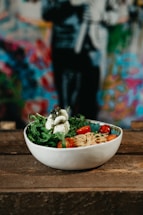 A bowl filled with pasta, fresh arugula, cherry tomatoes, and a serving of soft cheese sits on a wooden surface. The background is a vibrant, blurred mural with various colors.