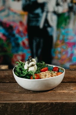 A bowl filled with pasta, fresh arugula, cherry tomatoes, and a serving of soft cheese sits on a wooden surface. The background is a vibrant, blurred mural with various colors.