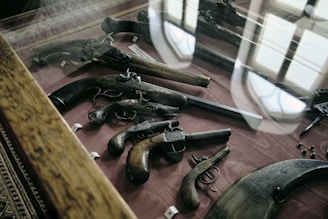 A display case showcasing various antique firearms is presented, including pistols and revolvers with intricate designs and wooden grips. The case is set on a dark red fabric and surrounded by a wooden frame. Reflections from overhead lighting and windows are visible on the glass surface of the case.
