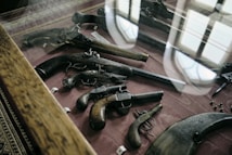 A display case showcasing various antique firearms is presented, including pistols and revolvers with intricate designs and wooden grips. The case is set on a dark red fabric and surrounded by a wooden frame. Reflections from overhead lighting and windows are visible on the glass surface of the case.