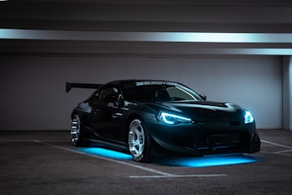 A sleek, modified sports car with aggressive styling sits in a dimly lit, empty parking garage. The car has bright blue LED lights highlighting its underside and headlights, giving it a futuristic appearance. It features a large rear wing and shiny alloy wheels, emphasizing its performance look.